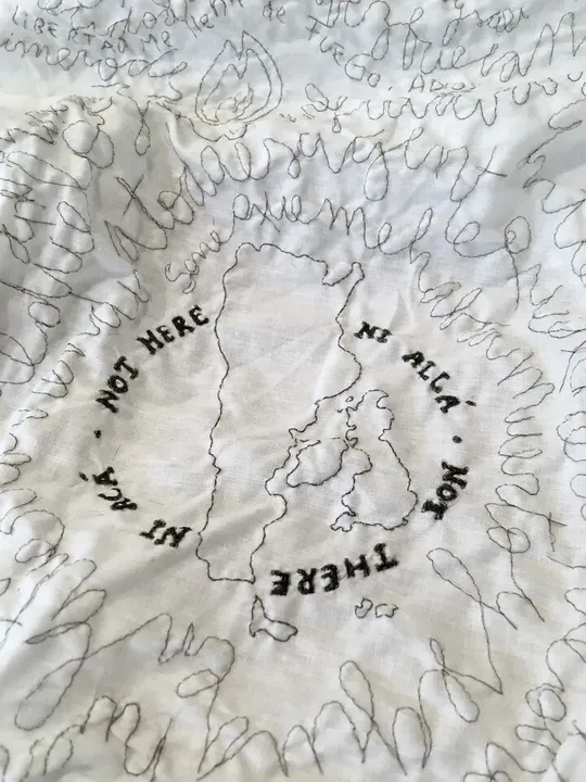 Detalle de la sábana bordada con pelo humano. Perteneciente a la obra Migran-t, el bordado muestra el mapa de Argentina junto al mapa de Reino Unido con la leyenda "Ni aquí ni allá" en inglés y español.